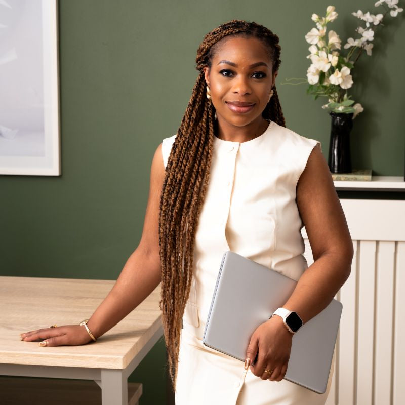 A woman with long braids stands indoors wearing a sleeveless cream dress and holding a silver laptop. She is posed next to a light wood table against a dark green wall, with a vase of white flowers in the background.
